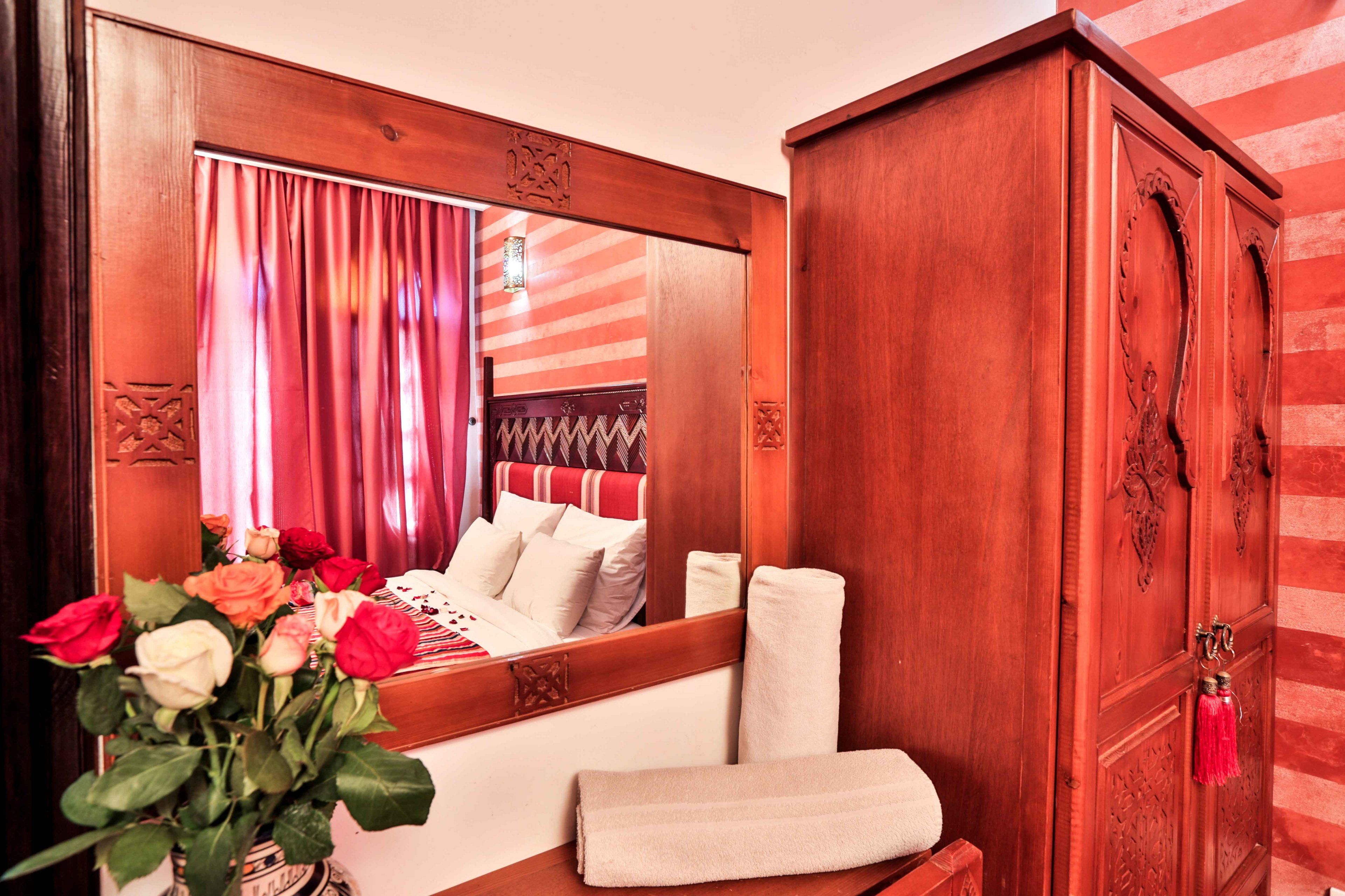 Habitación de hotel con un espejo grande, una cama con ropa blanca, un armario de madera y un florero con flores.