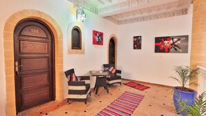 A lobby with a large wooden door marked '105', two wicker chairs, a small table, colorful rugs, and a potted plant. - Riad Nakhla (Essaouira)