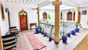 A covered courtyard with a tiled floor, potted plants, a wooden door, and a white railing. - Riad Nakhla (Essaouira)