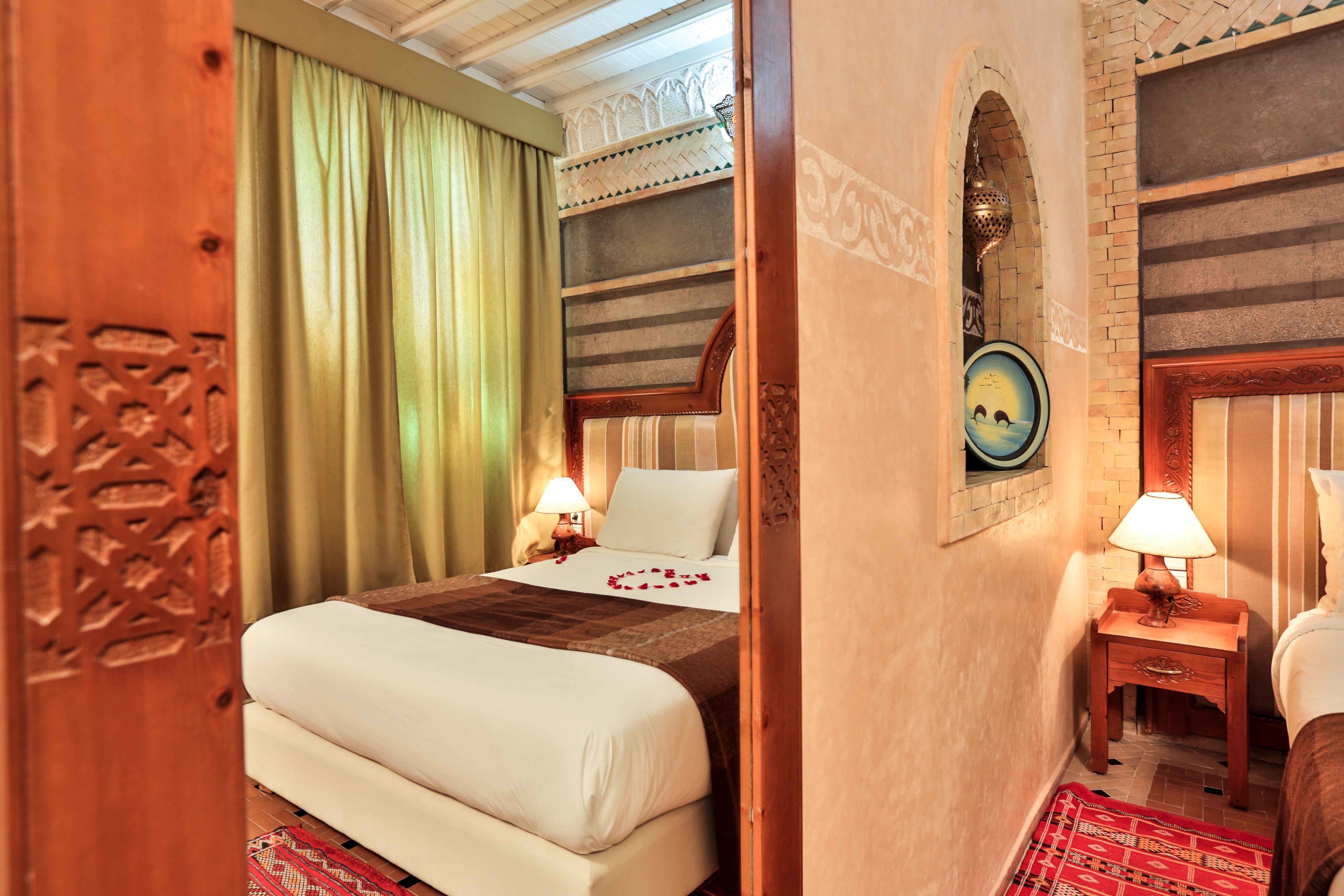 Una habitación de hotel con cama, mesita de noche con lámpara, pared con un cuadro y ventana con cortinas.