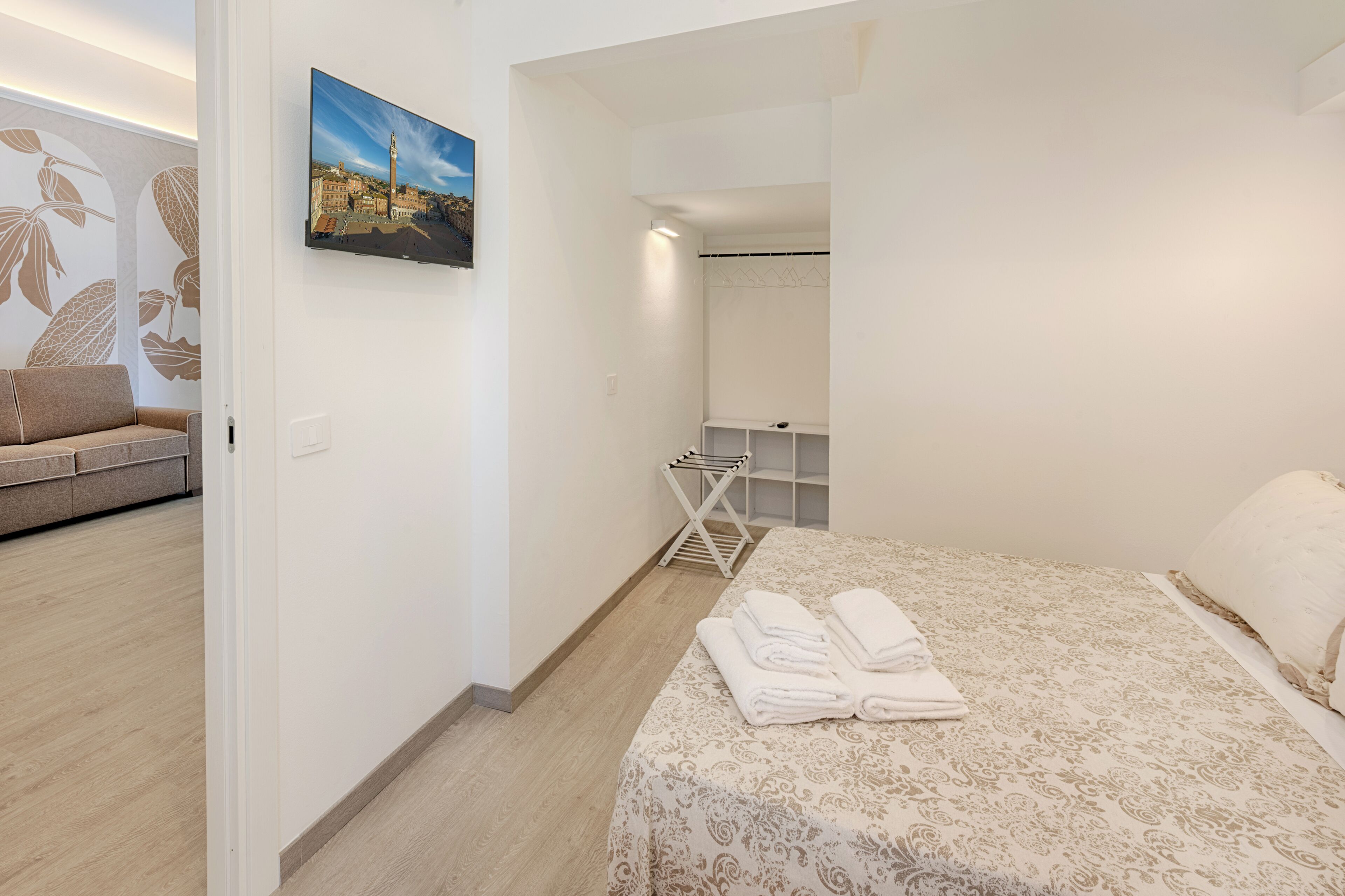 A hotel room with a bed, a seating area with a sofa, a wall-mounted TV, a drying rack, and a wardrobe.