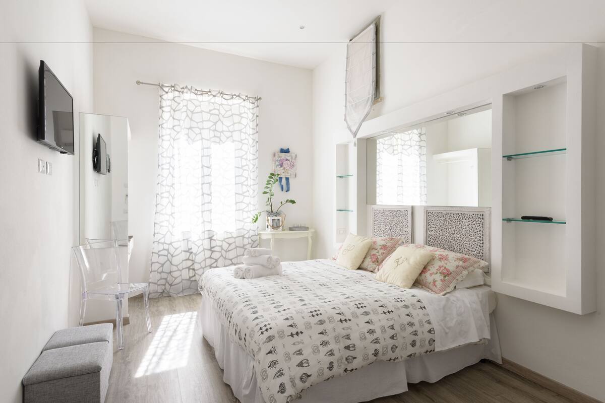 Ein modernes Schlafzimmer mit einem großen Bett, einem an der Wand montierten Fernseher und einem kleinen Beistelltisch mit einer Pflanze.