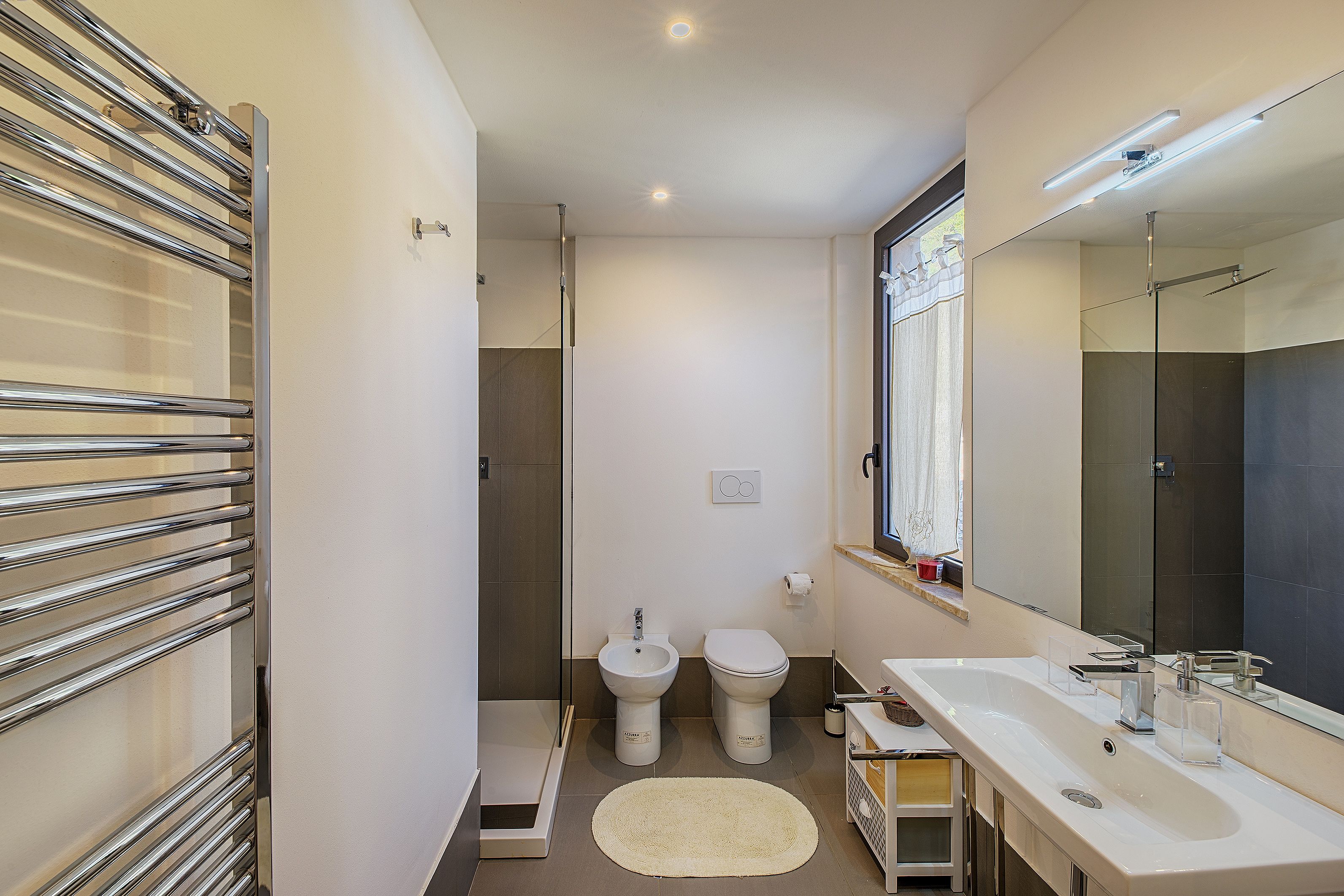 Une salle de bain moderne dotée d’une douche à l’italienne, d’un bidet, de toilettes, d’un grand miroir et d’un meuble-lavabo avec un évier.