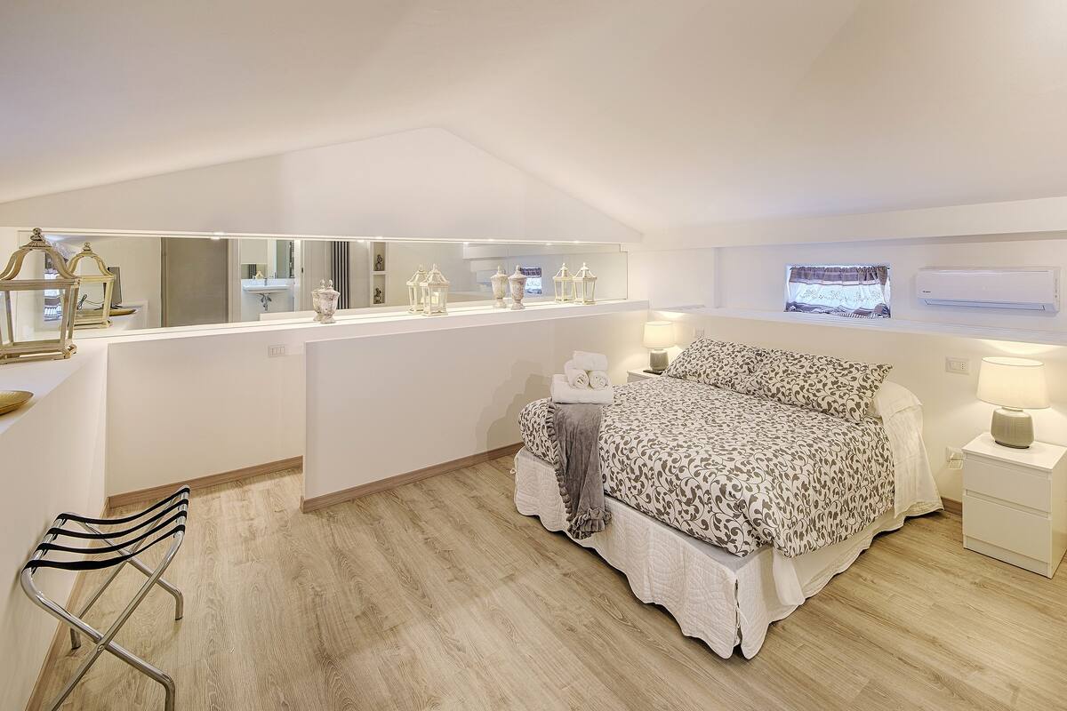 Ein Schlafzimmer mit einem großen Bett, einem Schminktisch mit Spiegel, einem Sessel und einem Fenster mit Vorhängen.