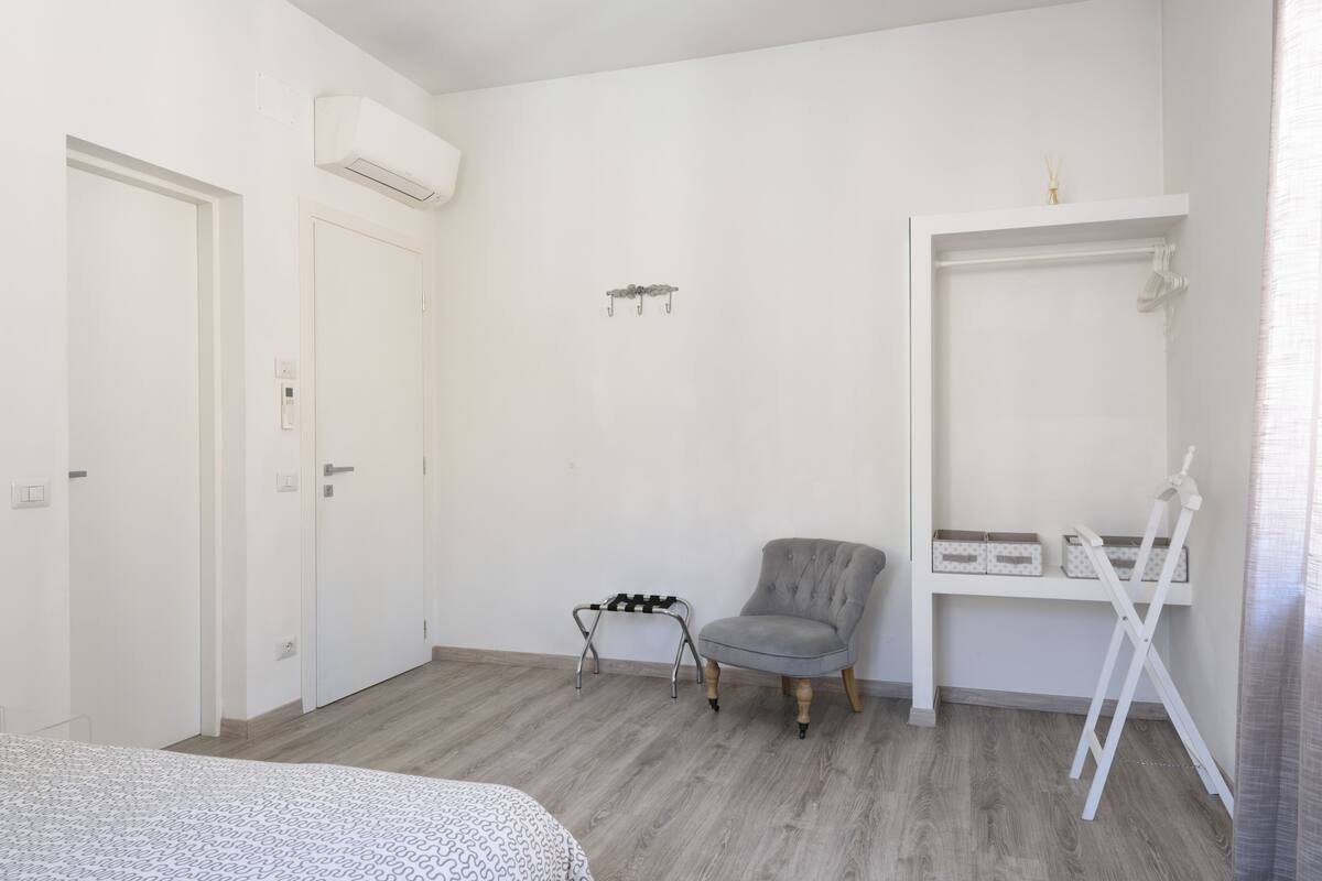 Ein Schlafzimmer mit einem Bett, einem grauen Sessel, einem weißen Wandschrank und einem weißen Regal mit Aufbewahrungsboxen.