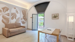 A modern living room with a sofa, a wooden dining table with chairs, a glass door leading outside, and a wall with floral artwork.
