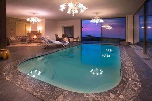 A modern indoor swimming pool with a glass bottom, surrounded by a stone-tiled floor, a fireplace, and a seating area with a view of the sunset. - Tenuta Bricchi (Sinio)