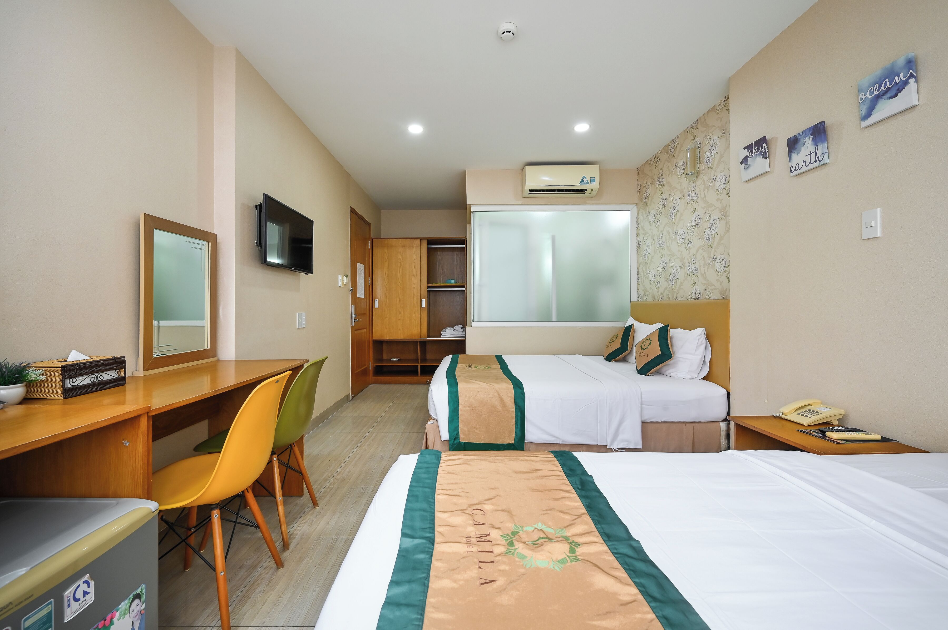 Habitación de hotel con escritorio, dos sillas, cama con edredón verde y blanco, televisor, aire acondicionado y ventana con vistas.