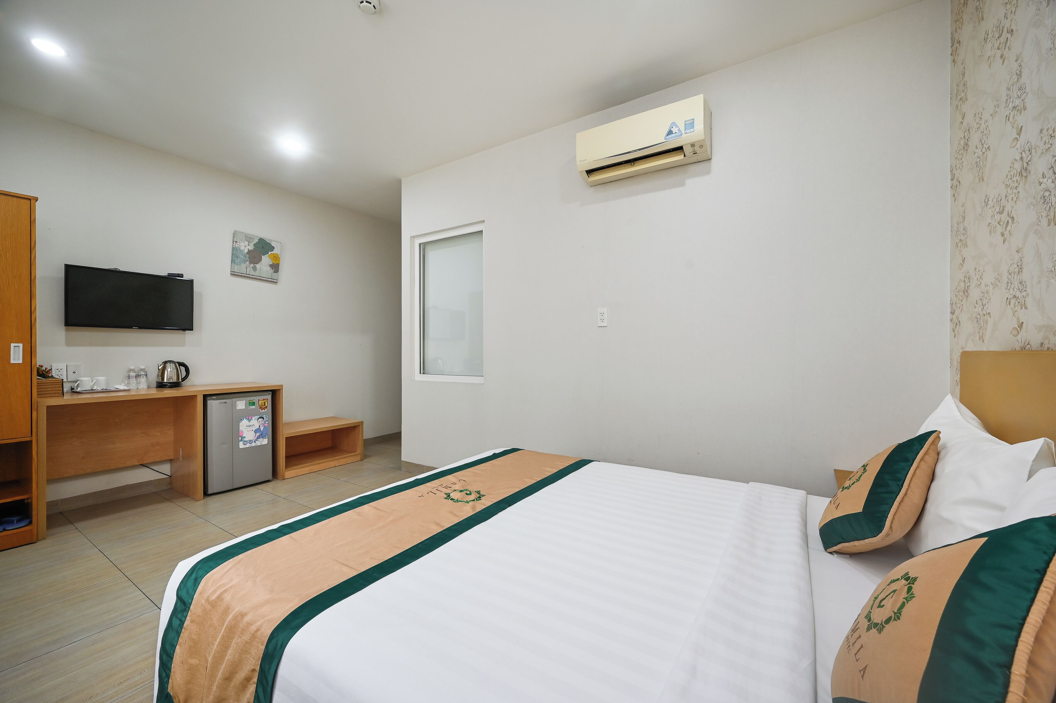 A hotel room with a bed, a desk with a refrigerator, a television, and an air conditioning unit.