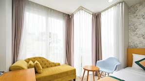 A hotel room with a yellow sofa, a blue chair, a bed with white bedding, and sheer curtains.