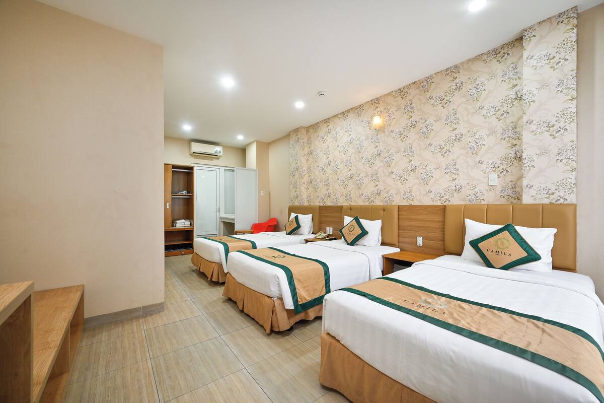 A hotel room with three beds, a wooden bench, a wardrobe, and a bathroom with a shower.