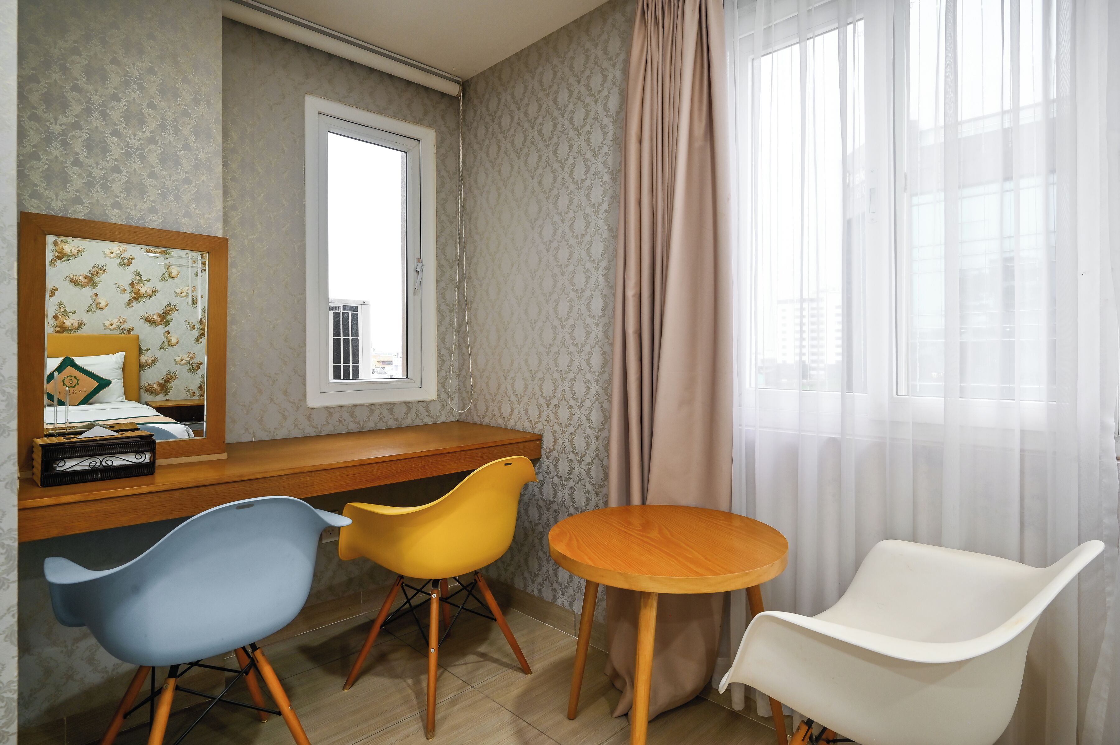 A room with a wooden desk, two chairs, a round table, a mirror, and a window with curtains.