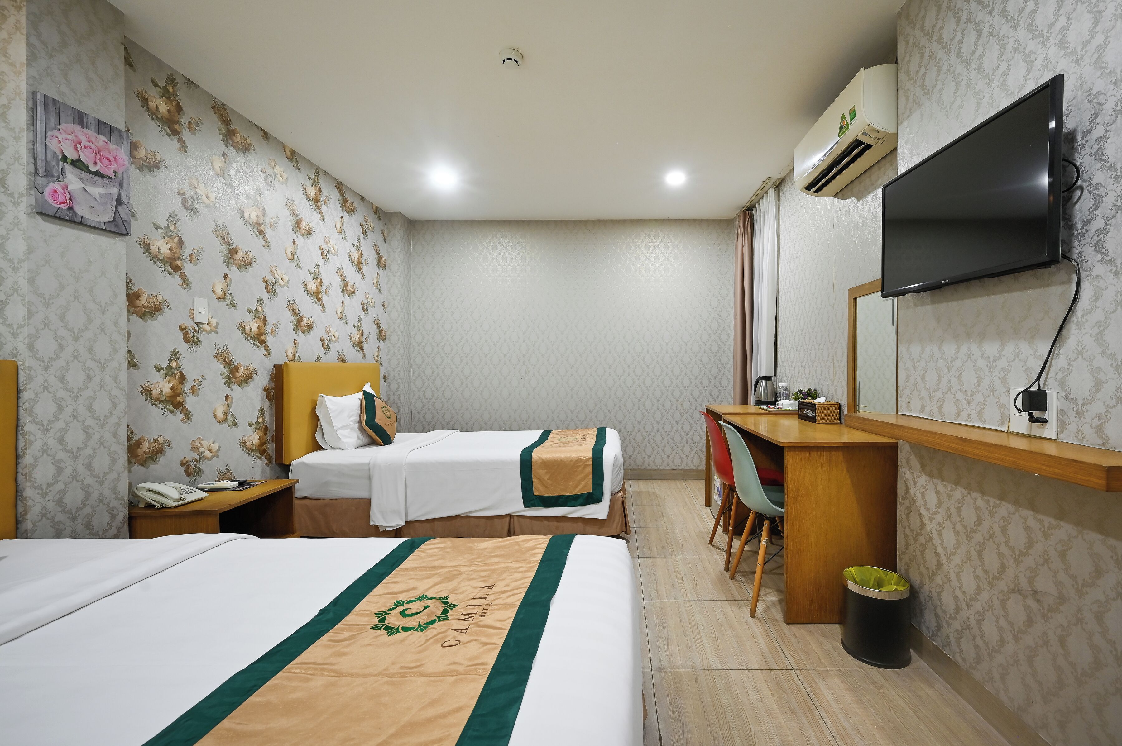A hotel room with two beds, a desk, a chair, a TV, and an air conditioning unit.