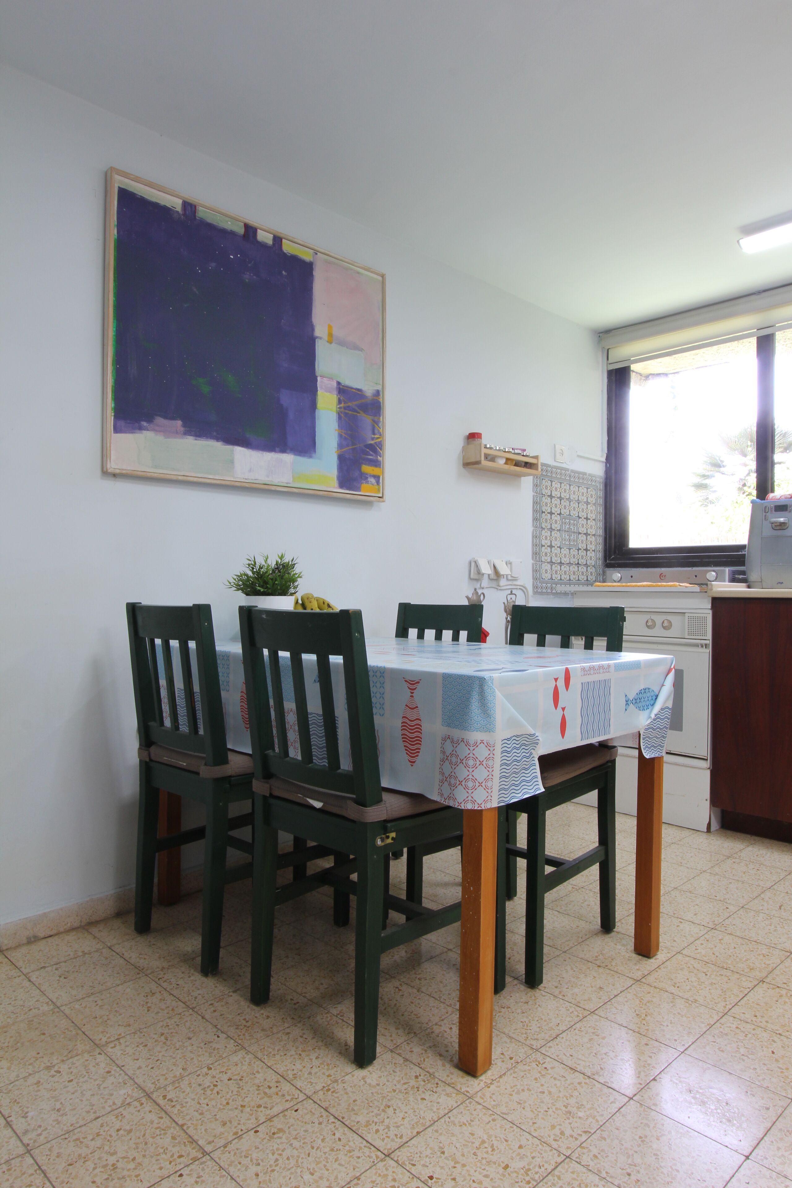 Zona pranzo con tavolo apparecchiato per quattro persone, sedie verdi, un quadro al muro e una finestra con persiane.