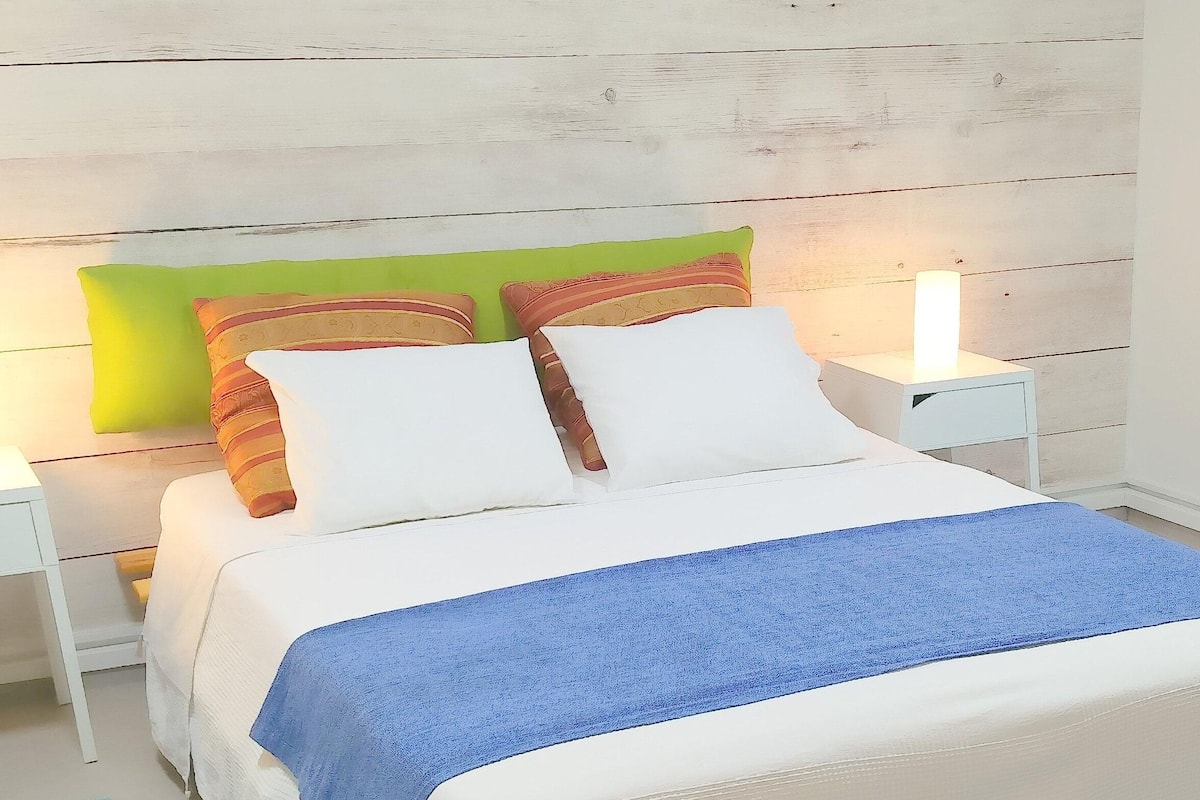 Ein ordentlich bezogenes Bett mit weißer und blauer Bettwäsche, zwei Nachttischlampen und bunten Kissen.