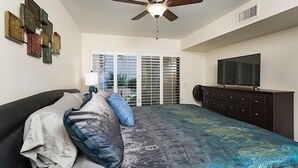 Une chambre à coucher comprenant un lit, un ventilateur de plafond, une télévision, une commode et une fenêtre avec des stores.
