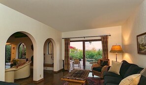 A living room with a sofa, armchair, and a patterned rug. There are two archways leading to an outdoor area with a view of greenery and a red umbrella. - Peak of Perfection By Signature Vacation Rentals (Tucson)