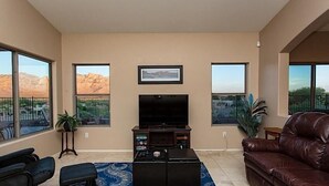 A living room with a large TV, two windows with mountain views, a brown leather couch, and a small table with a plant. - Scenic Spectacular By Signature Vacation Rentals (Oro Valley)