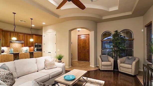 A spacious living area with a sofa, two armchairs, a coffee table, and a ceiling fan. A kitchen with wooden cabinets and a microwave is visible in the background. - Sail Away By Signature Vacation Rentals (Chandler)