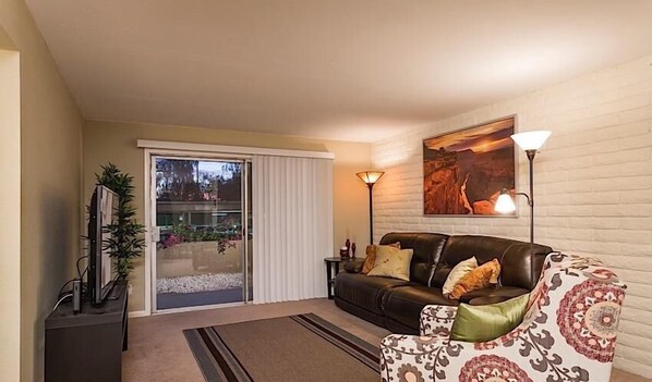 A living room with a sliding glass door, a black leather sofa, a patterned armchair, a floor lamp, and a painting on the wall.