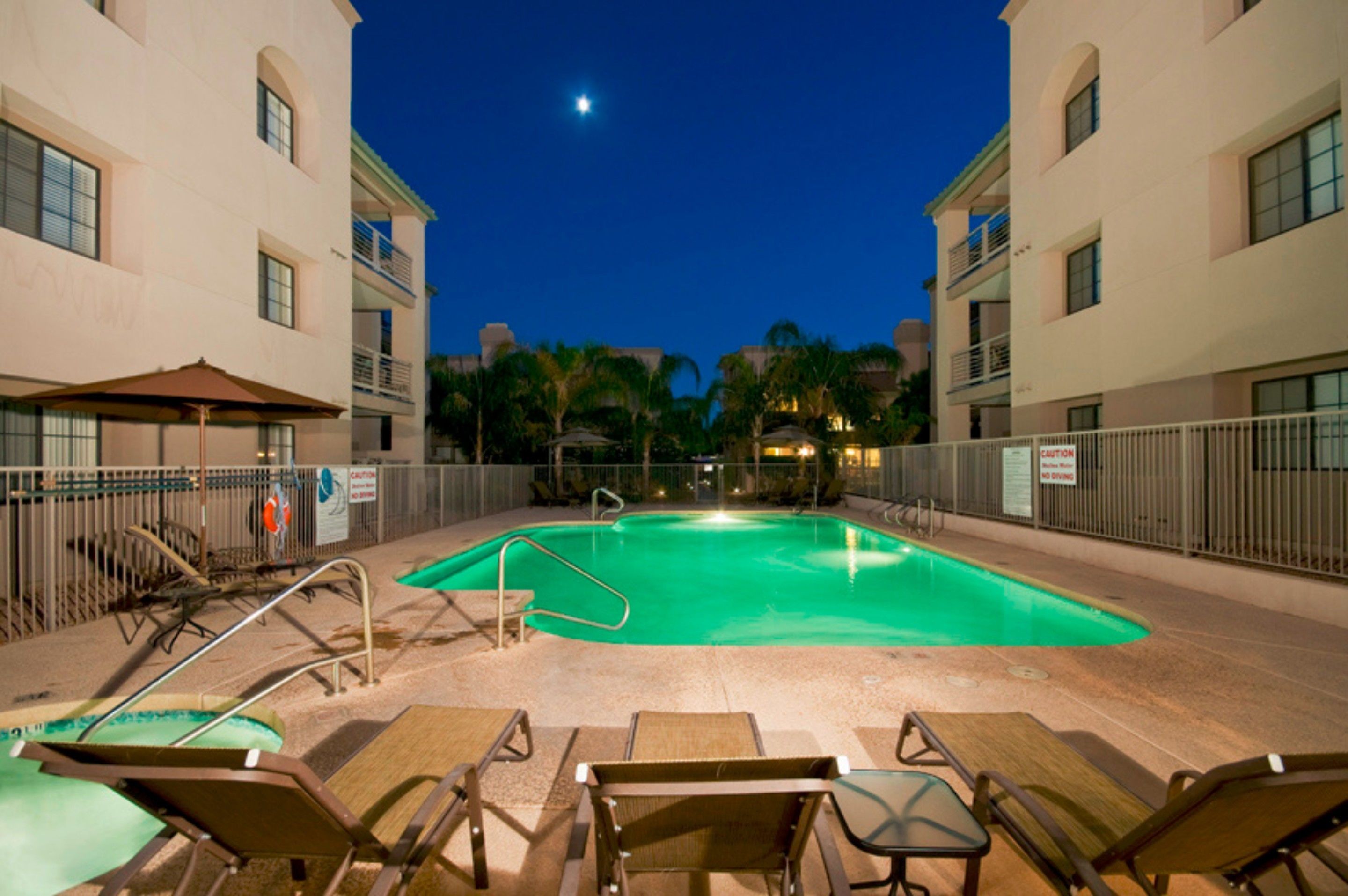 A swimming pool with lounge chairs and umbrellas, surrounded by a fence and palm trees.