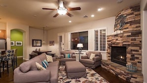 A living room with a stone fireplace, a ceiling fan, and a dining area with a green wall and a table. - Palm Valley By Signature Vacation Rentals (Goodyear)