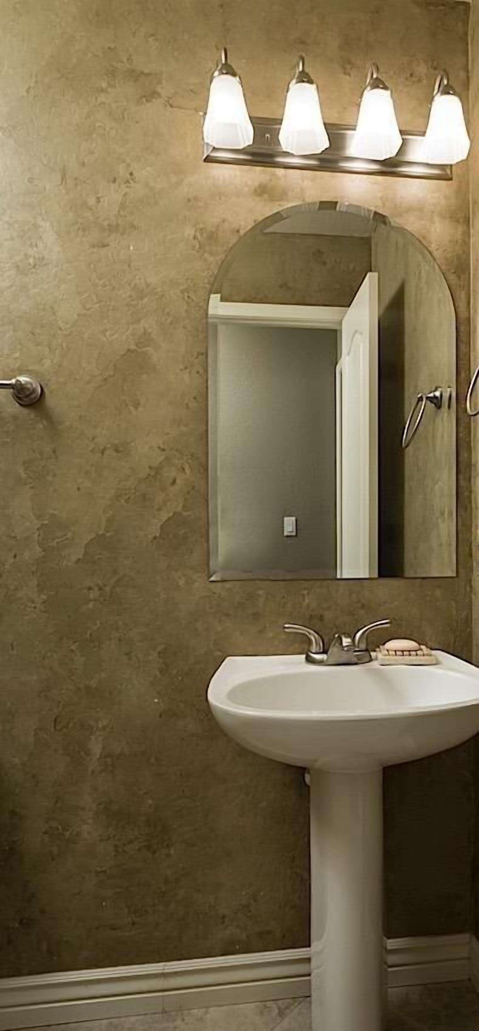 Une salle de bain avec un lavabo sur colonne blanc, trois appliques murales et un miroir au-dessus du lavabo.