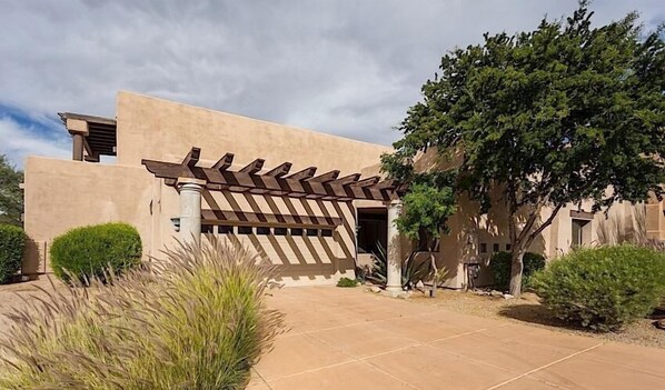 A modern building with a unique pergola entrance, surrounded by landscaped gardens and a clear sky.