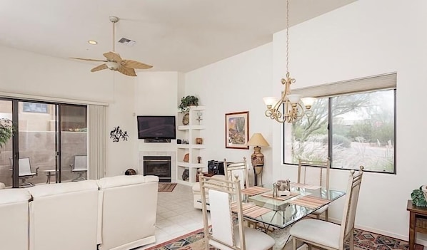 A spacious living room with a glass dining table, white sofas, a ceiling fan, a fireplace, and a large window offering a view of the outdoors.