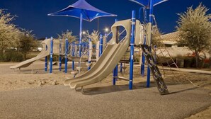 A playground with slides and climbing structures, surrounded by trees and a building in the background.