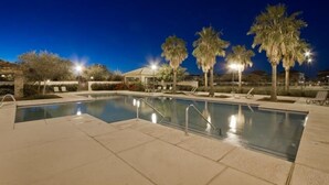 An outdoor swimming pool at night with palm trees and lounge chairs.
