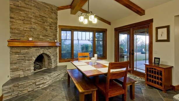 A dining area with a stone fireplace, wooden beams, large windows, and a view of the outdoors. - Majestic Carefree Home By Signature Vacation Rentals (Carefree)