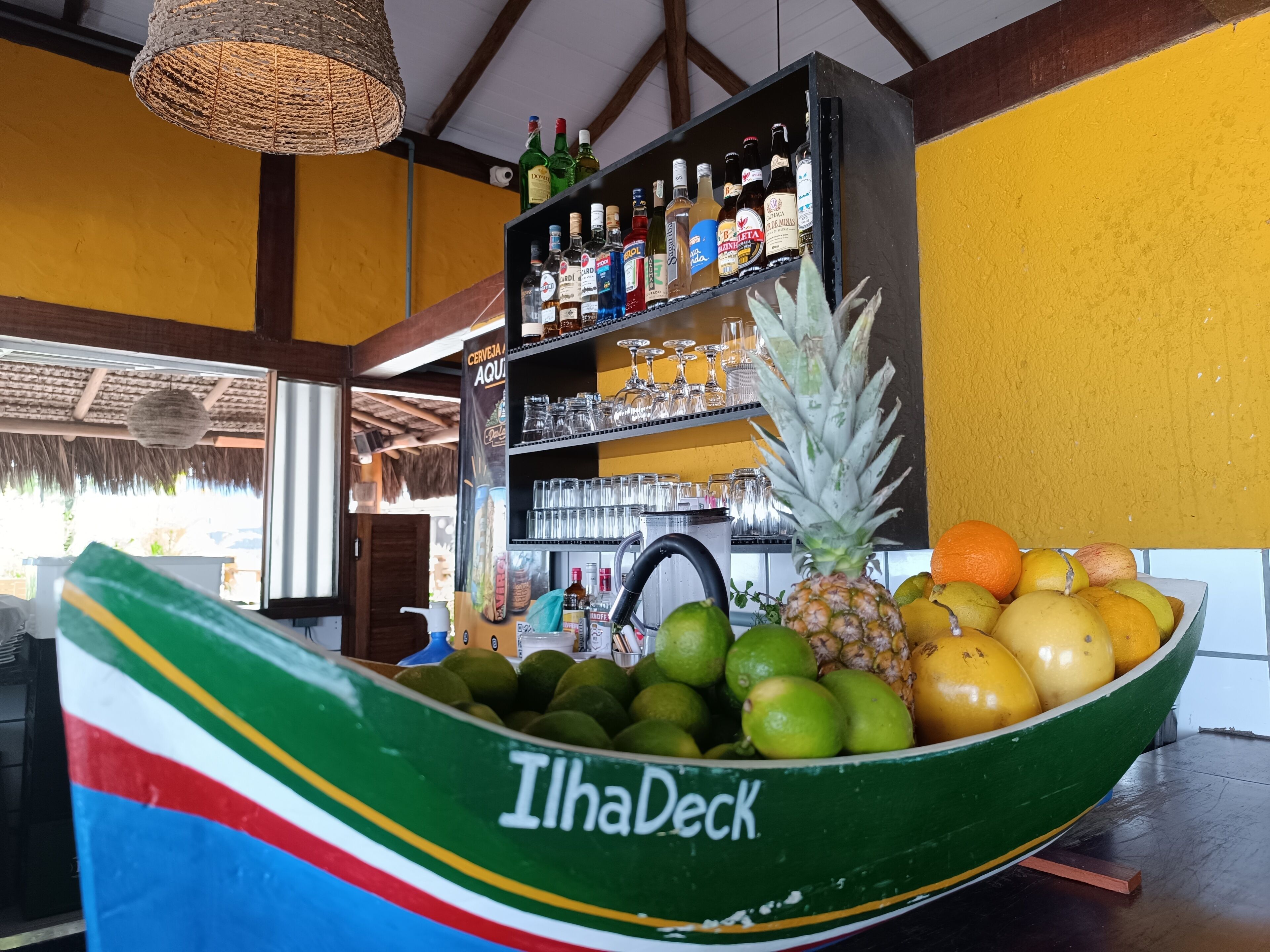 Un coin bar avec une étagère remplie de bouteilles d’alcool, un ananas et un saladier de fruits.