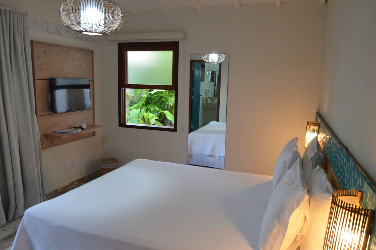 Hotelkamer met een bed, een televisie op een houten plank, een raam met uitzicht op groen en een spiegel die een deel van de kamer weerspiegelt.