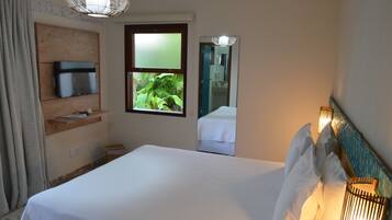 Quarto de hotel com uma cama, uma televisão sobre uma prateleira de madeira, uma janela com vista para vegetação e um espelho refletindo parte do quarto.