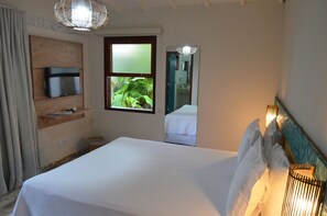 Habitación de hotel con cama, televisor en un estante de madera, ventana con vista a follaje y un espejo que refleja parte de la habitación.