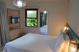 Habitación de hotel con cama, televisor en un estante de madera, ventana con vista a follaje y un espejo que refleja parte de la habitación.