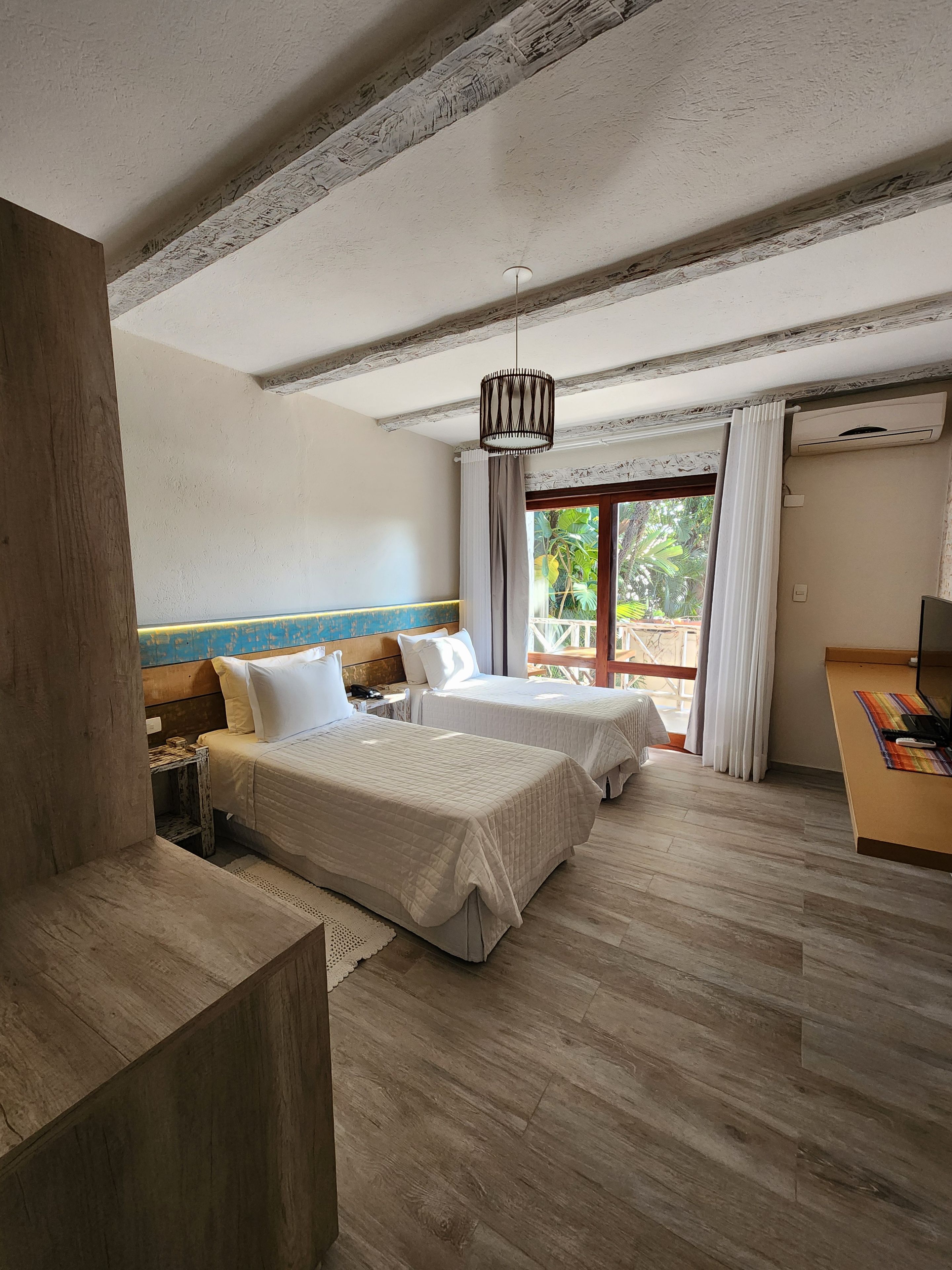 Habitación de hotel con dos camas, techo de madera, ventilador de techo, ventana con cortinas y una pequeña mesa con lámpara.