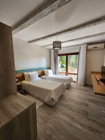 Habitación de hotel con dos camas, techo de madera, ventilador de techo, ventana con cortinas y una pequeña mesa con lámpara.