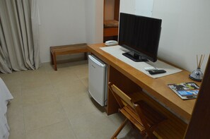 A small, tidy room with a wooden desk, a computer, a small refrigerator, a bench, and a bed with a brown blanket.