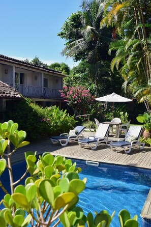 Un área de piscina con sillones de descanso y una mesa bajo una pérgola, rodeada de vegetación exuberante y palmeras.