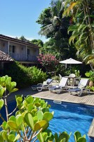 Un área de piscina con sillones de descanso y una mesa bajo una pérgola, rodeada de vegetación exuberante y palmeras.