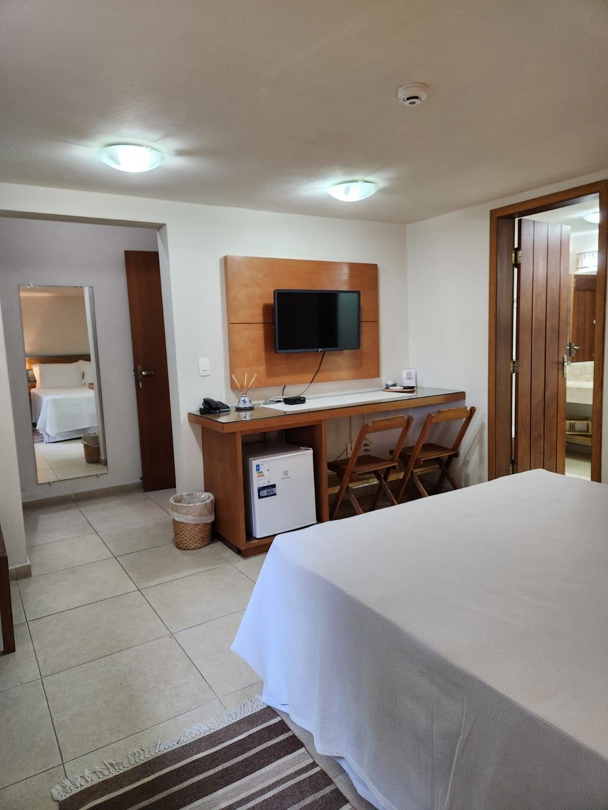 Quarto de hotel com cama, escrivaninha, televisão de tela plana, cadeira, pequeno refrigerador e lixeira.