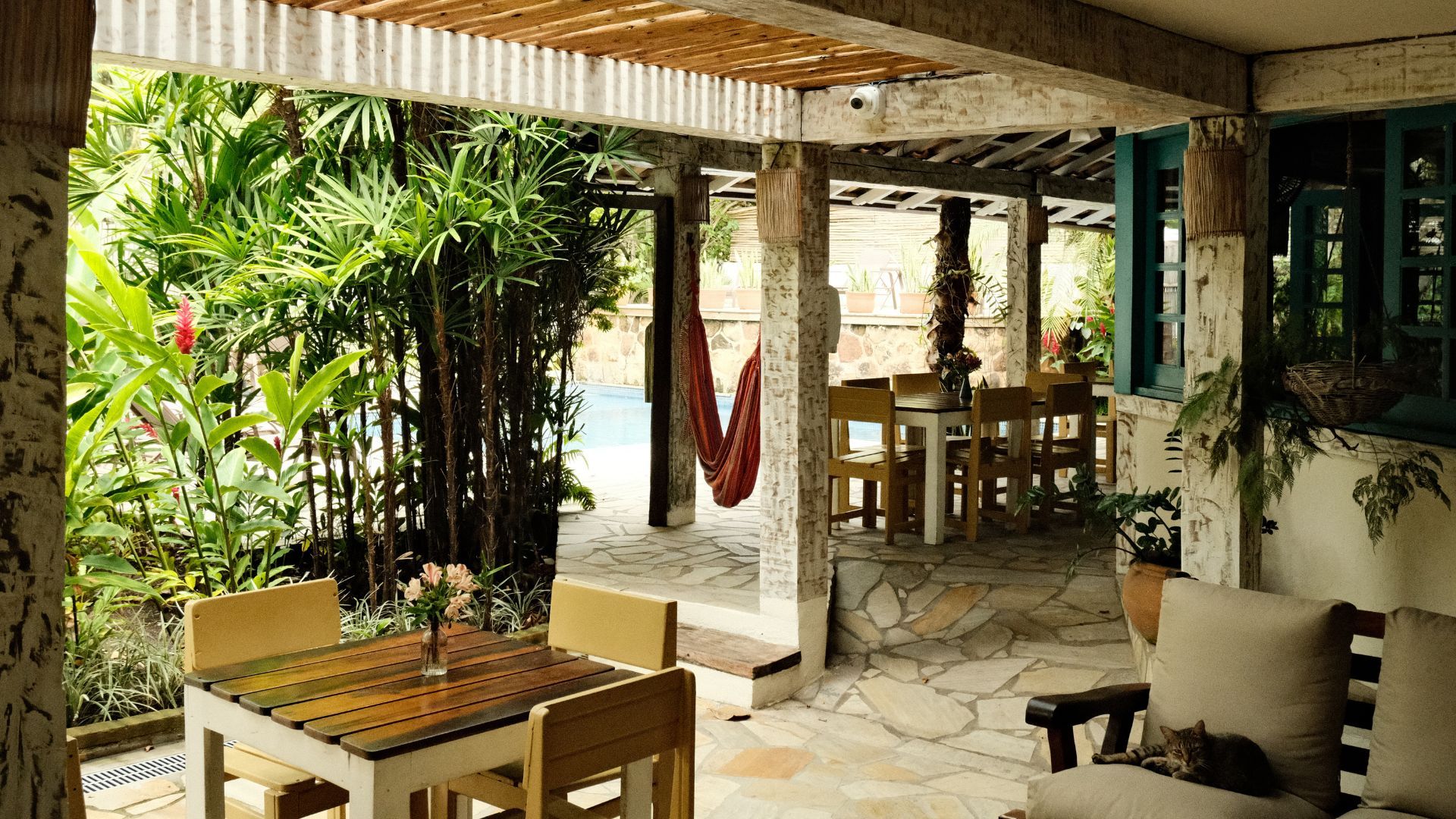 Área de estar externa com piscina, móveis de madeira e plantas tropicais exuberantes.