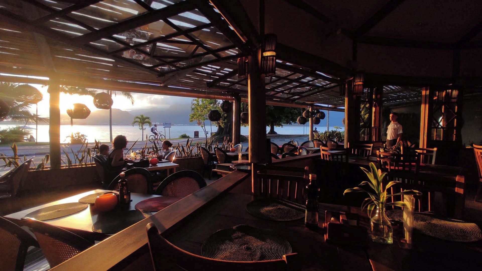 Un restaurante al aire libre con mesas y sillas, vista al mar y una persona de pie junto a la ventana.