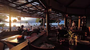 Un restaurante al aire libre con mesas y sillas, vista al mar y una persona de pie junto a la ventana.