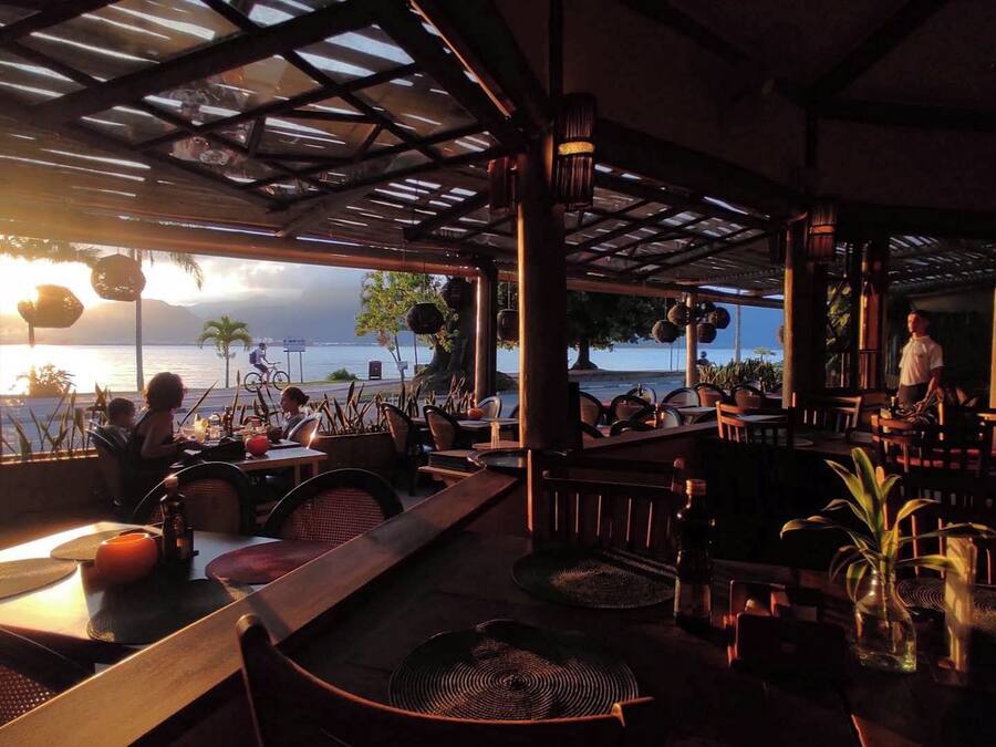 An outdoor restaurant with tables and chairs, a view of the sea, and a person standing by the window.