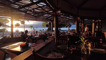 Restaurante ao ar livre com mesas e cadeiras, vista para o mar e uma pessoa parada perto da janela.