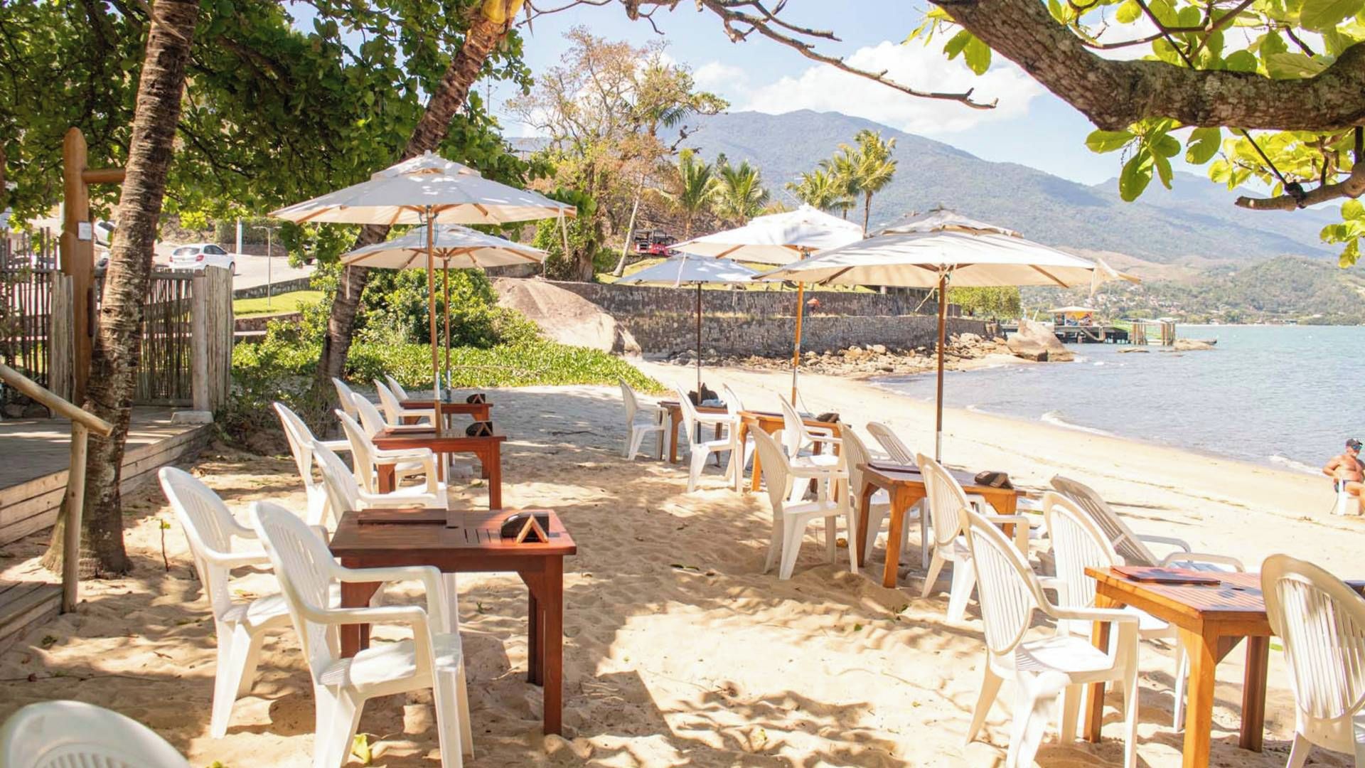A beachside dining area with tables and chairs, umbrellas, and a view of the sea and mountains.