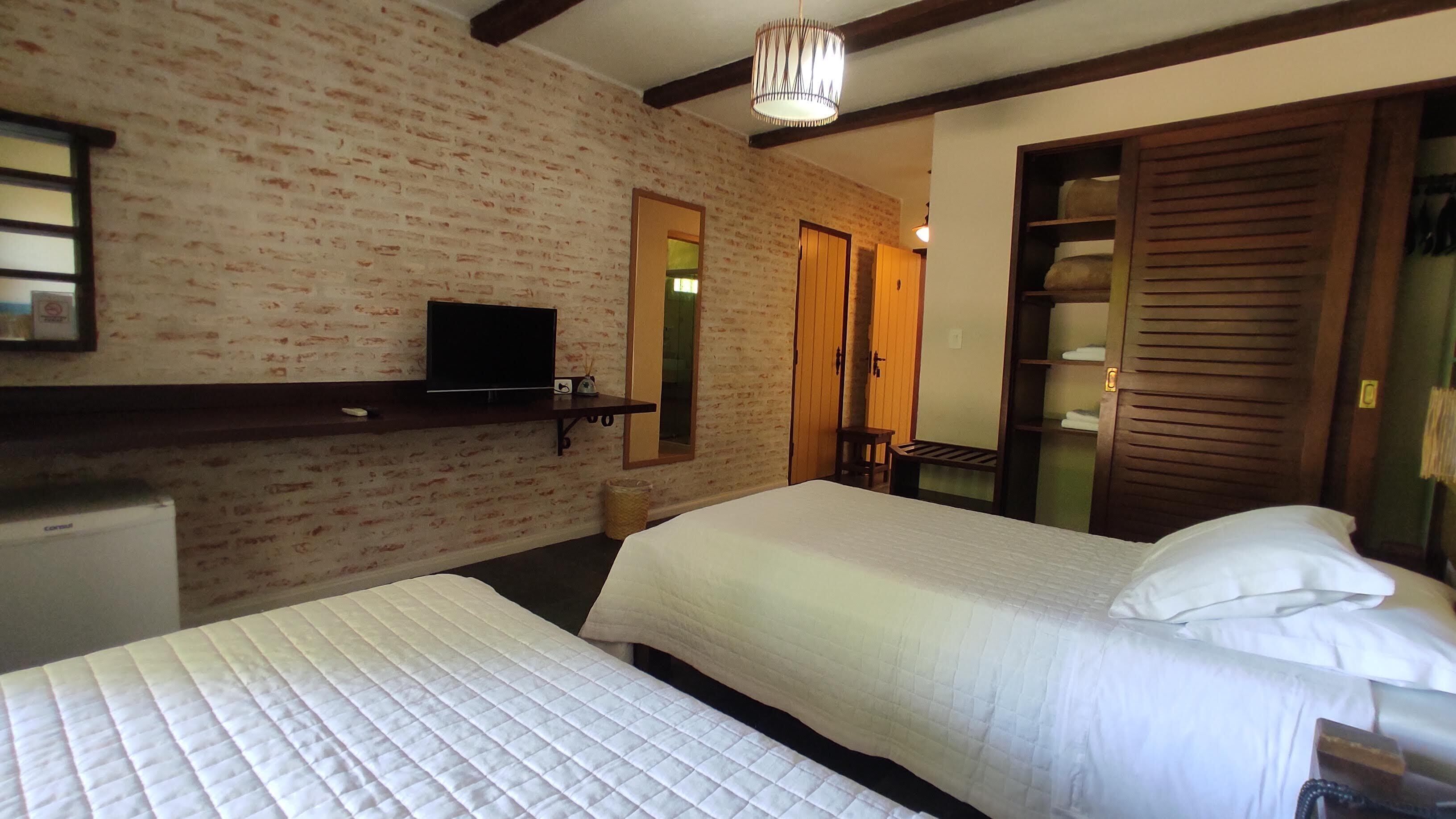Una habitación de hotel con dos camas, un televisor en un estante, un armario de madera y una pared de ladrillo.